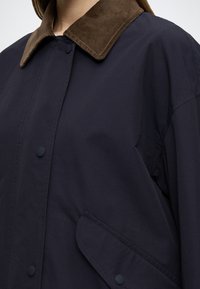 Námornícky kabát s hnedým kordurovým golierom, cvokmi a bočným vreckom. Má hladkú textúru a uvoľnený strih.