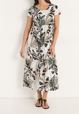 Femme portant une robe midi à manches courtes avec un motif de feuilles tropicales vert, noir et beige et des sandales noires à bout ouvert.