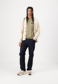 Beige knitted cardigan with large buttons, olive green T-shirt featuring a logo, dark jeans, and beige sneakers with black accents.
