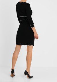 Svart klänning med trekvartslånga ärmar, scalloperad vit kant vid midjan och fållen, texturerat tyg, matchad med svarta högklackade skor.