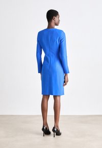 Ένα εφαρμοστό μπλε φόρεμα με μακριά μανίκια, περιλαμβάνοντας στρογγυλή λαιμόκοψη και φερμουάρ στην πλάτη, συνδυασμένο με μαύρα τακούνια.