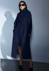 Marineblauwe wollen trenchcoat met een hoge kraag, een ceintuur in de taille en zijzakken. Glad textuur, lange mouwen en een lengte tot halverwege de kuit.