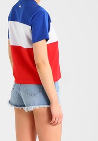 Person som bär en trikolor tröja i blått, vitt och rött med korta ärmar samt ljusblå fransade jeansshorts, visad bakifrån.