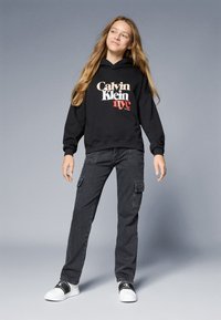 Čierna mikina s kapucňou s logom "Calvin Klein nyc" v bielej a červenej farbe. V kombinácii s tmavými cargo nohavicami a čiernymi nazúvacími topánkami s logovými akcentmi.
