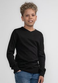 Schwarzer, langärmeliger V-Ausschnitt-Pullover aus weichem Material, mit gerippten Bündchen und Saum, kombiniert mit blauen Jeans. Das Model posiert mit den Händen in den Taschen.