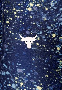 Witte stierenkop logo op blauwe stof met verspreide gele en lichtblauwe verfspetterpatroon en zichtbare stiksels.