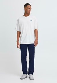 En ung man står mot en ljus bakgrund, klädd i en stor vit t-shirt, marinblå byxor och grå sneakers, med händerna avslappnat vid sidorna.