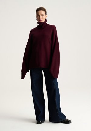 Kobieta stojąca, ubrana w za duży bordowy sweter z golfem, szerokie ciemnoniebieskie dżinsy i czarne buty na jednolitym tle.