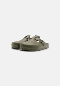 Kaki klomper laget av glatt plast, med en justerbar stropp og en teksturert såle med grep for bedre feste. Rund, bred tådesign.