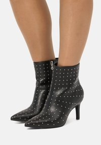 Botas negras hasta el tobillo, hechas de cuero brillante, con punta afilada y adornadas con pequeños tachuelas plateadas. Cuentan con una cremallera lateral y un tacón delgado.