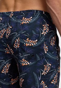 Maritime svømmetights med blomsterdesign i oransje og hvitt, med elastisk midjebånd og en baklomme med svart knappe detalj.