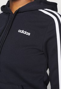 Sudadera con capucha azul marino con diseño de tres rayas blancas en las mangas, cierre de cremallera y logo bordado de Adidas en el pecho. Textura de tela suave.