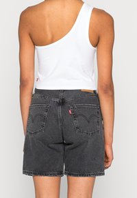 Crop top bianco a spalla unica abbinato a pantaloni shorts in denim grigio scuro, con tasche posteriori distintive e una texture del tessuto scolorita. Dettaglio del logo rosso visibile.
