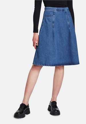 Model trägt einen knielangen blauen Denim A-Linien-Rock mit Fronttaschen, ein schwarzes langärmliges Top und schwarze klobige Plateau-Schuhe.
