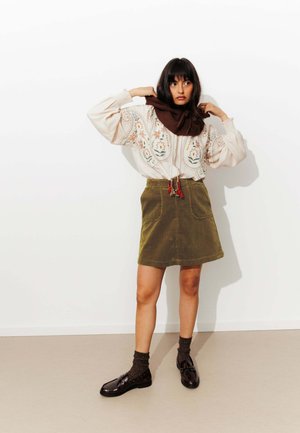 Femme ajustant une écharpe marron, portant un blouse brodée de fleurs, une jupe en velours côtelé olive, des chaussettes noires et des mocassins noirs, debout contre un mur blanc.