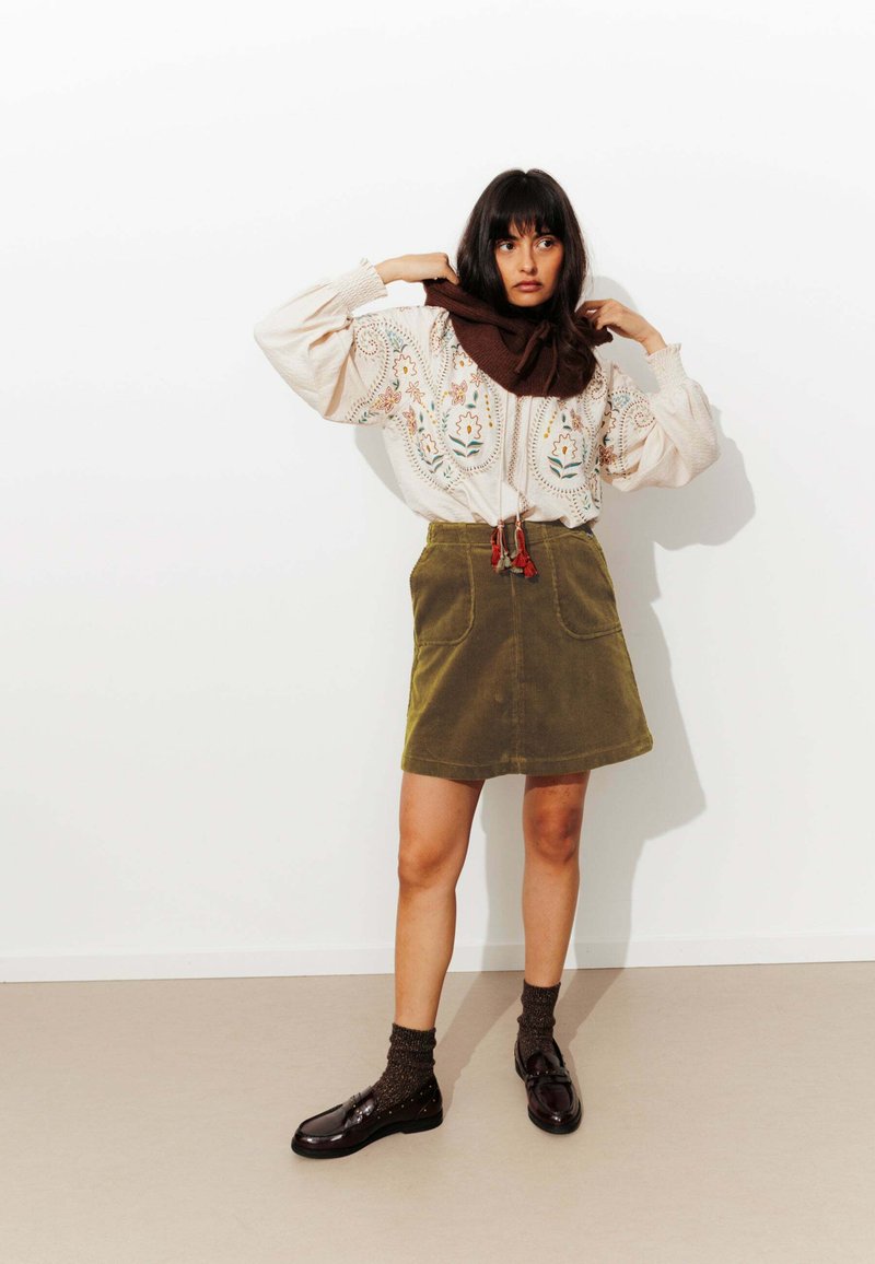 Femme ajustant une écharpe marron, portant un blouse brodée de fleurs, une jupe en velours côtelé olive, des chaussettes noires et des mocassins noirs, debout contre un mur blanc.