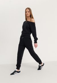 Sweatshirt noir décontracté à épaules dénudées avec une coupe ample, associé à un pantalon de jogging noir avec une taille élastique et des coutures latérales, complété par des chaussures de sport noires.