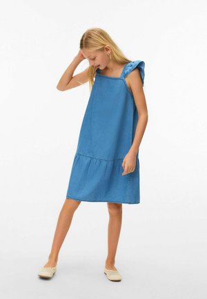 Fille en robe en jean bleue sans manches avec des bretelles à volants regardant vers le bas, debout avec une jambe pliée, portant des chaussures beige sans lacets.