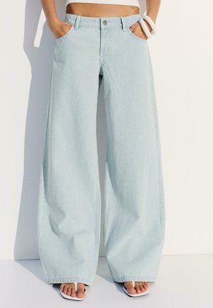 Osoba nosząca jasnoniebieskie, szerokie spodnie w pionowe paski i białe sandały, z rękami w kieszeniach, stojąca przy białej ścianie.