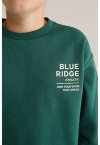 Zelena dukserica s rebrastim ovratnikom, sa bijelim tekstom: "BLUE RIDGE," "ATHLETIC," i "OWN YOUR GAME STAY AHEAD." Mješavina pamuka, ležeran dizajn.