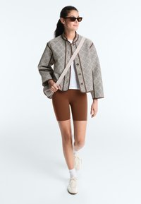 Femme marchant portant une veste à motifs, une chemise blanche, un short de cyclisme marron, des baskets blanches, des lunettes de soleil et un sac bandoulière.