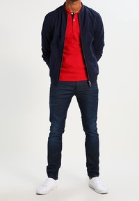 Marinblå bomberjacka över en röd polo, mörkblå jeans som sitter tajt, och vita sneakers. Klara färger och en snygg, enkel design.