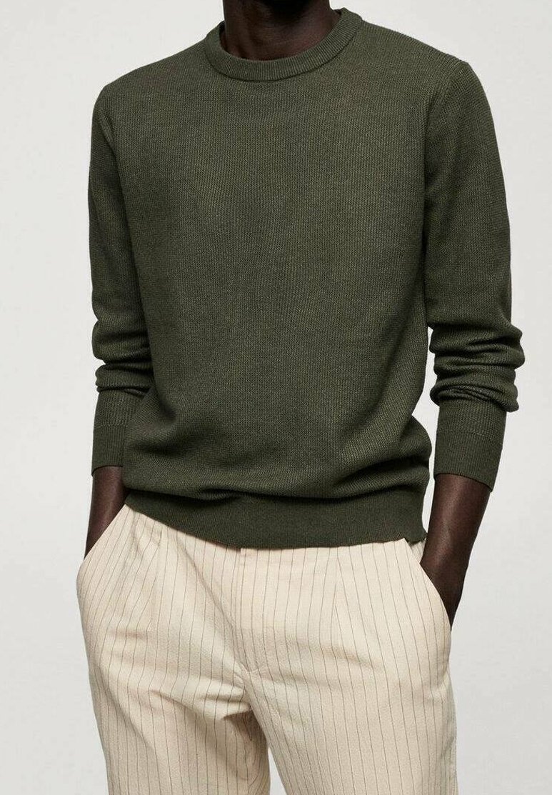 Personne portant un pull en tricot vert olive et un pantalon crème à fines rayures, les mains dans les poches, sur un fond uni.