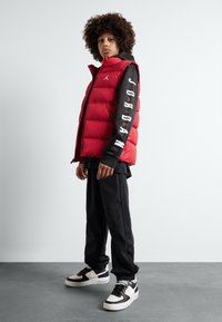Gilet rembourré rouge avec des accents noirs, présentant le texte « JORDAN » sur les manches. Associé à un pantalon noir et des baskets bicolores. Fond à surface plate.
