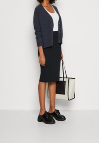 Cardigan navy con bottoni in legno, canotta bianca, gonna a tubino nera, scarpe nere con lacci, mentre tiene una grande borsa a tracolla bianca e nera.