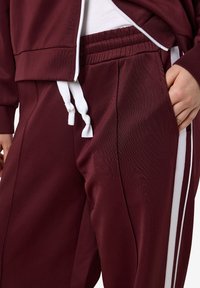 Lähellä otettu kuva henkilöstä, joka wears maroon-väriset verkkarit, joissa on valkoiset sivuraidat, valkoiset nyörit, elastinen vyötärö ja matching maroon-värinen takki vetoketjulla.