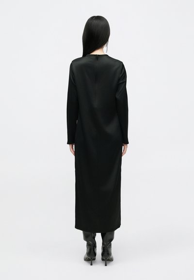 Vestido de cetim preto de mangas longas com silhueta reta, decote redondo e fecho na parte de trás. Usado com botas de salto alto pretas até ao joelho.