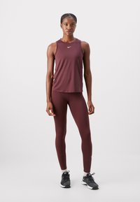 Maroon športna majica brez rokavov in legice, narejene iz elastičnega materiala. Nike logo na majici. Model nosi črne športne copate z belimi poudarki.