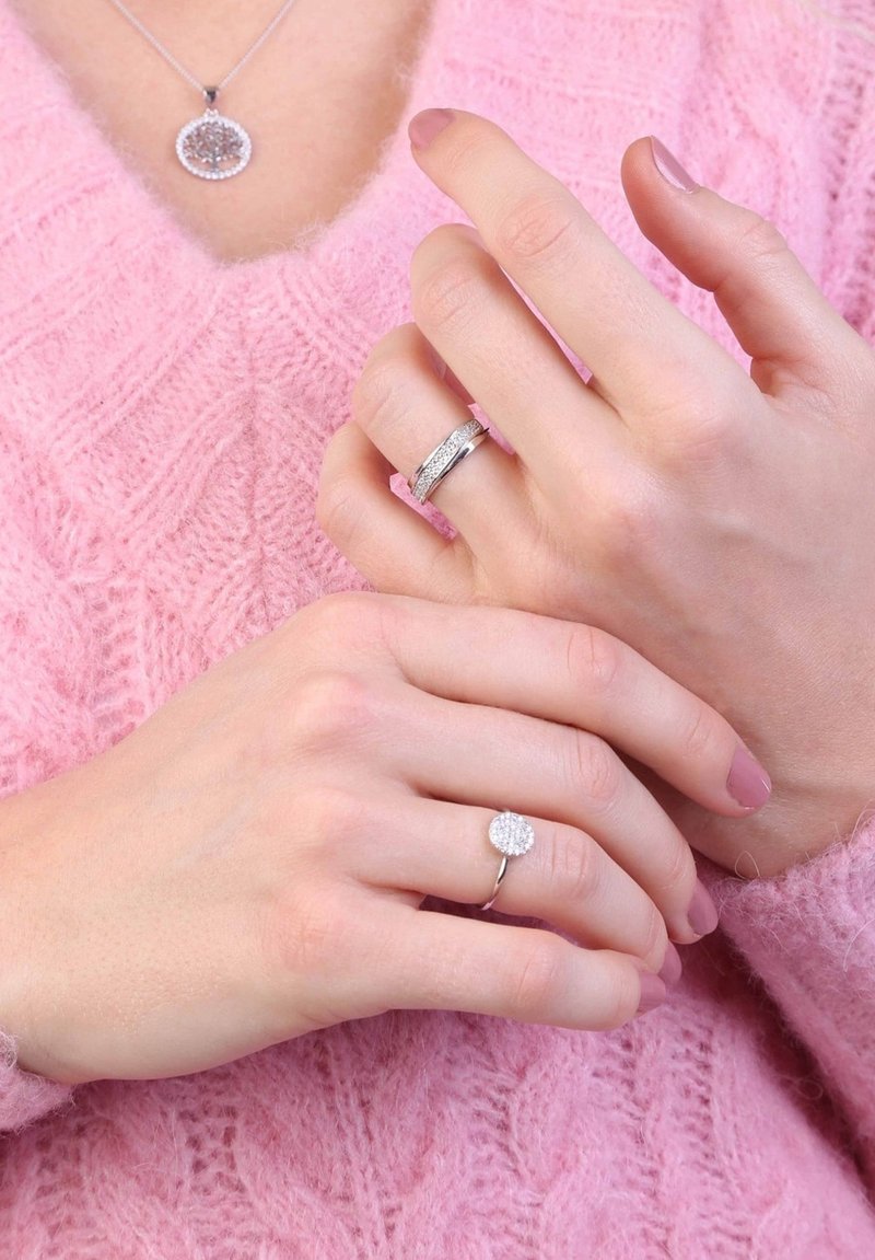 Silver jewelry features a round pendant necklace and two rings. The ring on the left has a band with textured detail; the right has a circular, paved design.