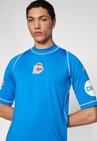 Modré sportovní tričko s kontrastními světle modrými švy. Na hrudi má odznak a na rukávech loga. Elastická, hladká tkanina.