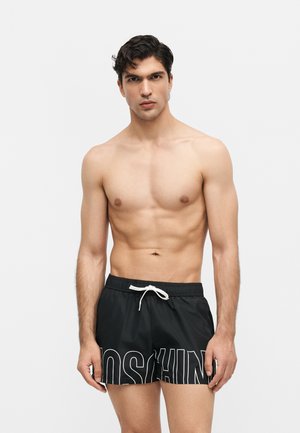 Ing nélküli férfi sötét hajjal, fekete zsinóros fürdőgatya, nagy fehér kontúros betűkkel egy egyszerű, világos háttér előtt.