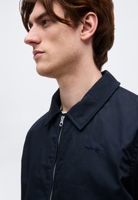 Jeune homme à la peau claire et aux cheveux bruns portant une veste zippée bleu marine foncé avec un col, tourné vers la gauche sur un fond uni.