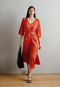 Rochie luminoasă din satin roșu, cu mâneci scurte, detaliu cu nod la talie și fustă vaporoasă, asociată cu tocuri negre cu bretele și o geantă tote neagră.