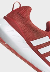 Röd sportskor med övre i stickad textil, vit detalj med tre ränder, texturerad häl och tjock vit sula med röd accent.