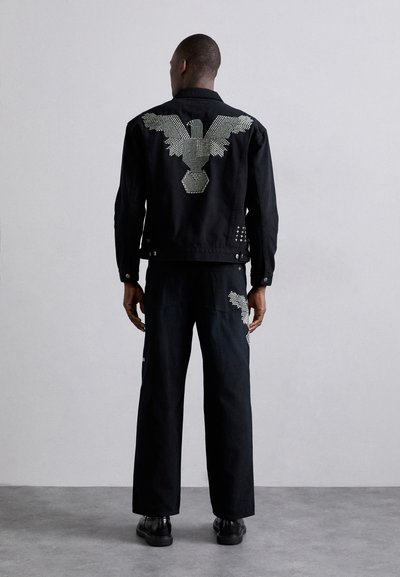 Chaqueta de mezclilla negra con diseño de águila de strass en la parte de atrás, combinada con pantalones anchos negros que presentan un patrón de acento.