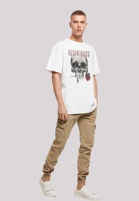 White cotton t-shirt with a skull and rose graphic, beige cargo pants, and white sneakers, standing on a neutral background.