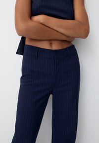 Personne portant un pantalon bleu marine à rayures fines et un haut sans manches assorti, les bras croisés sur le ventre nu.
