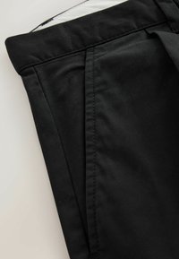 Nahaufnahme von schwarzen Stoffhosen mit Bund und Seitentasche mit sichtbarer Naht auf hellem Hintergrund.