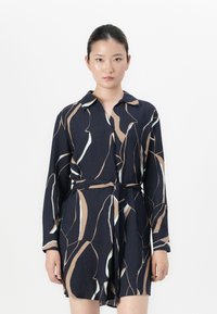 Femme portant une robe noire à manches longues cintrée avec des motifs abstraits beige et blancs, se tenant devant un fond blanc uni.