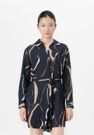 Femme portant une robe noire à manches longues cintrée avec des motifs abstraits beige et blancs, se tenant devant un fond blanc uni.