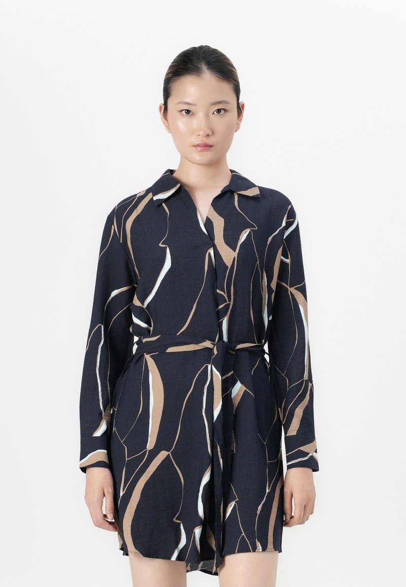 Femme portant une robe noire à manches longues cintrée avec des motifs abstraits beige et blancs, se tenant devant un fond blanc uni.