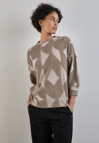 Beige chlupatý svetr s diamantovým vzorem v krémové barvě. Má kulatý výstřih a volné rukávy, nošený s černými kalhotami.