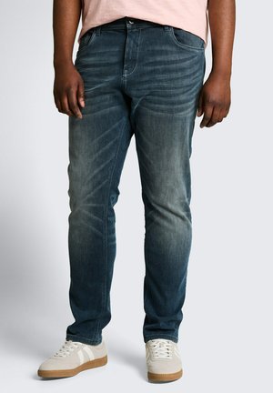 Mann trägt ausgewaschene blaue Jeans, hellrosa Hemd und weiße Sneakers mit Gummisohlen, steht vor einem schlichten grauen Hintergrund.