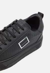 Baskets noires en cuir texturé présentant un motif crocodile, des lacets ronds et une plaque logo rectangulaire argentée sur le côté.