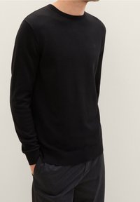 Maglione nero in maglia con scollo rotondo, polsini e orlo a coste. Presenta un logo sottile sul petto. Texture liscia, design aderente.