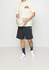 Krémská košile s krátkým rukávem a růžovou grafikou na hrudi, v kombinaci s černými sportovními šortkami, modrými ponožkami a bílými atletickými botami, držící basketbal.